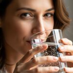 A beautiful woman with natural makeup and a slight smile, looking directly at the camera while holding a glass of clear water, ready to drink. The image is bright and shows a natural detoxification method. This image illustrates the simple steps in debunking detox myths for a healthier you.