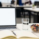 A glass of water and a bowl of berries and nuts sit next to a laptop and notebook on a clean, modern office desk, with a blurred background of a busy office.