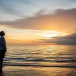A person stands on a calm beach at sunset, looking out at the horizon. The sky shows a mix of clearing storm clouds and warm light, symbolizing peace after a period of struggle with anxiety.