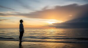 A person stands on a calm beach at sunset, looking out at the horizon. The sky shows a mix of clearing storm clouds and warm light, symbolizing peace after a period of struggle with anxiety.