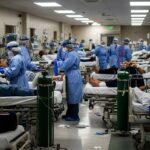 A realistic illustration showing a hospital emergency room during a pandemic, with exhausted medical staff in PPE tending to numerous patients on gurneys and beds. The scene is crowded and chaotic under harsh fluorescent lighting.