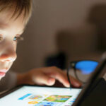 A child looks at a tablet, with a pair of glasses on the table nearby.
