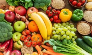 A vibrant, overhead shot showcases a diverse selection of fresh, whole foods arranged on a wooden surface. Colorful fruits like red and green apples, oranges, and yellow bananas mingle with bunches of green grapes. An assortment of vegetables includes green broccoli florets, red, yellow, and green bell peppers, ripe red cherry tomatoes on the vine, and slender cucumbers. Small wooden bowls contain various grains and seeds. The overall impression is one of abundance, health, and the natural goodness of unprocessed ingredients.