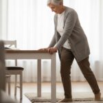 A realistic illustration of a senior woman at home, holding onto a table for support while carefully stepping over a small rug. The setting is a bright, clean living room.