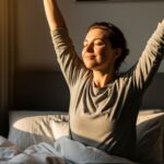 A person is sitting up in bed in a sunlit room, looking refreshed and stretching their arms above their head, symbolizing the mental clarity and peak performance that comes from better sleep.