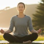 A woman sits cross-legged on a yoga mat in a grassy area near the beach, with her eyes closed and a serene expression on her face. The background features a green hill and the ocean, with soft sunlight from the sunrise illuminating the scene. Her relaxed posture and open hands on her knees suggest a practice of meditation or mindfulness.