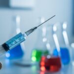A gloved hand holds a syringe filled with blue liquid in a laboratory setting with scientific equipment and a DNA helix model in the background.