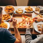 Two people seen from behind and above, excessively eating a variety of unhealthy foods including pizza, pasta, fried chicken, and cakes.