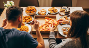 Two people seen from behind and above, excessively eating a variety of unhealthy foods including pizza, pasta, fried chicken, and cakes.