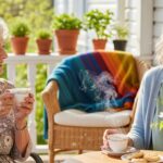 Two cheerful elderly women sitting on a porch, laughing and drinking tea.