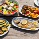 A vibrant flat lay of a Mediterranean outdoor lunch with Greek salad, grilled fish, roasted vegetables, and hummus, representing healthy diet plans.
