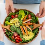 A top-down, close-up photo of two pair of hands tossing a colorful and fresh salad in a wooden bowl, featuring grilled chicken, avocado, and tomatoes on a clean kitchen counter.