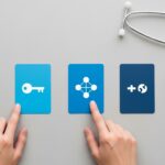 Hands pointing to three blue cards with icons representing health insurance options (key, network, plus symbol) next to a stethoscope, symbolizing the decision-making process for health plans like HMO, PPO, and EPO