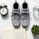Flat lay of gray running shoes, black alarm clock, glass of water, and open book on a white marble table, illustrating a healthy morning routine for better sleep.