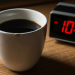A chipped white mug filled with black coffee next to a digital clock displaying 10:14 AM on a wooden table in low light.